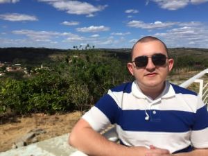 Sidney est&aacute; usando &oacute;culos escuros em frente a uma paisagem com &aacute;rvores bem verdes, c&eacute;u azul claro com nuvens e veste uma camisa listrada azul e branca (Foto: Sidney Andrade/Arquivo Pessoal)